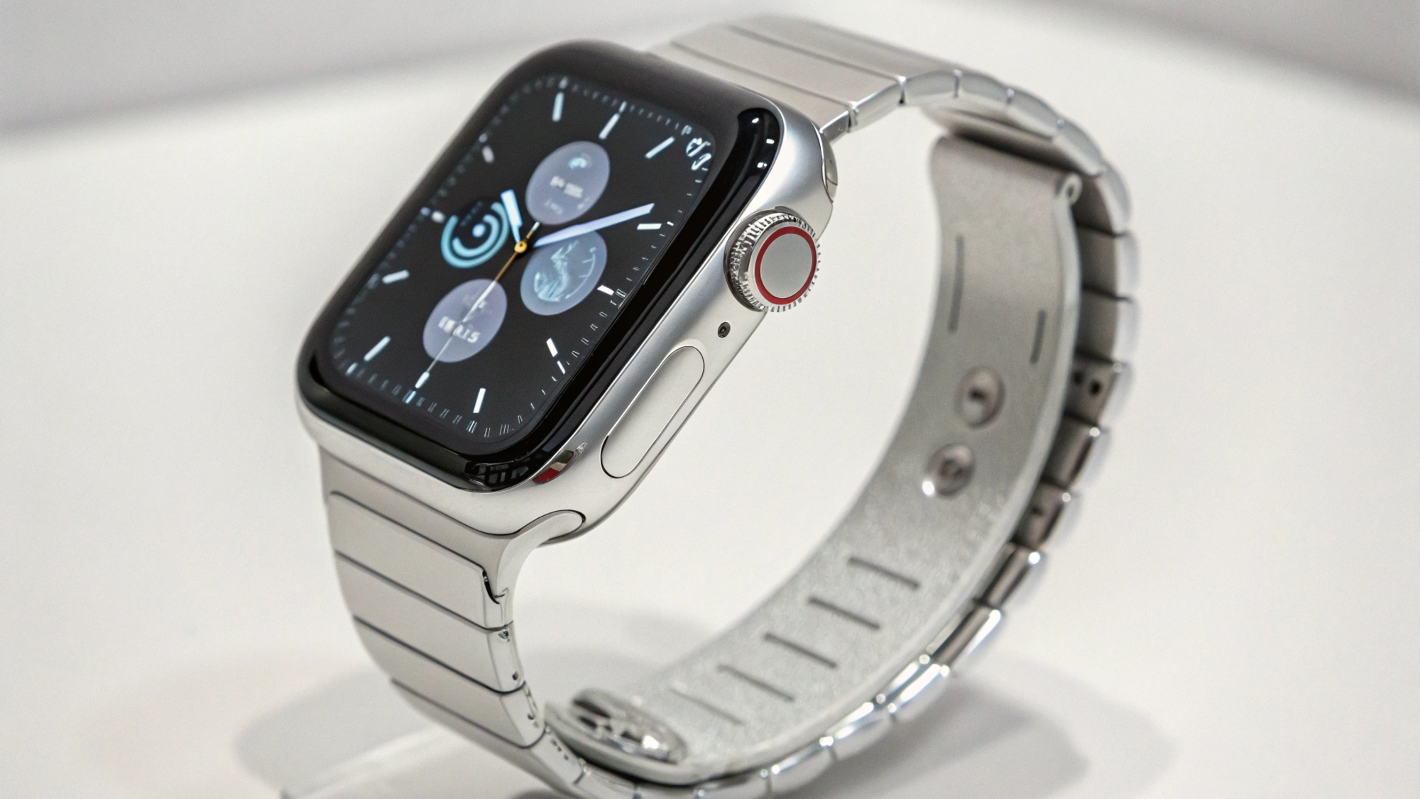 На снимке изображены серебристые умные часы Apple Watch с металлическим браслетом. Циферблат часов отображает текущее время, а также несколько круглых виджетов с различной информацией. Сбоку на корпусе часов видна заводная головка с красной деталью. Браслет состоит из отдельных звеньев и имеет матовое покрытие с внутренней стороны. Задняя часть часов также имеет матовое покрытие и детали с винтами. Часы слегка наклонены вперед и располагаются на белой поверхности.